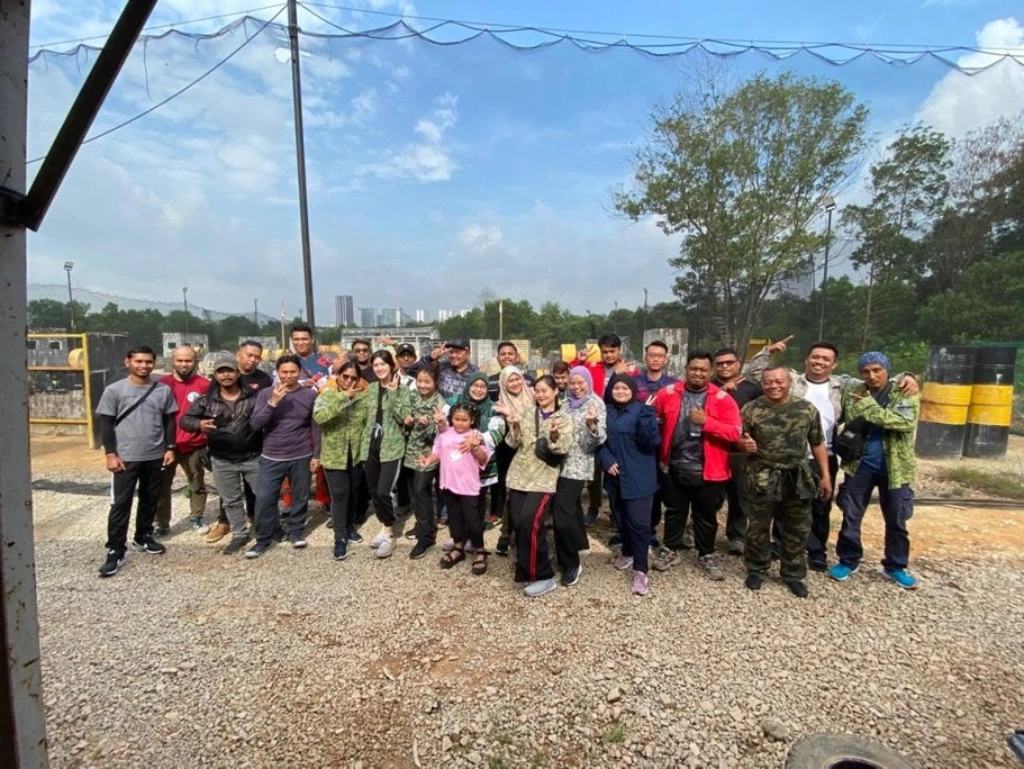 A large group of people posing outdoors at a paintball arena, with many wearing camouflage jackets over their casual clothes.