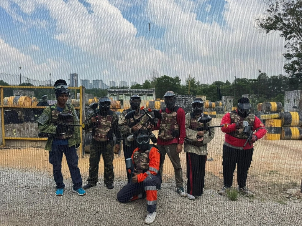 Seven people fully geared up in camouflage paintball uniforms and protective masks posing with their paintball markers on a gravel field.
