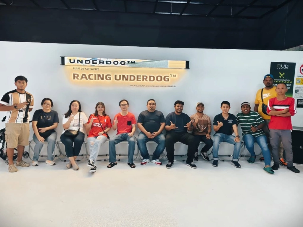 A group of eleven people sitting and standing together in front of an illuminated "RACING UNDERDOG" sign at an indoor go-kart facility.