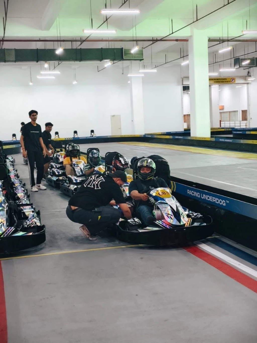 Drivers wearing helmets sitting in their go-karts on an indoor track while a staff member assists the person in the front kart.