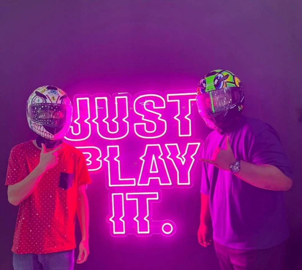 Two individuals wearing brightly patterned full-face helmets posing in front of a glowing pink neon sign that reads "JUST PLAY IT."