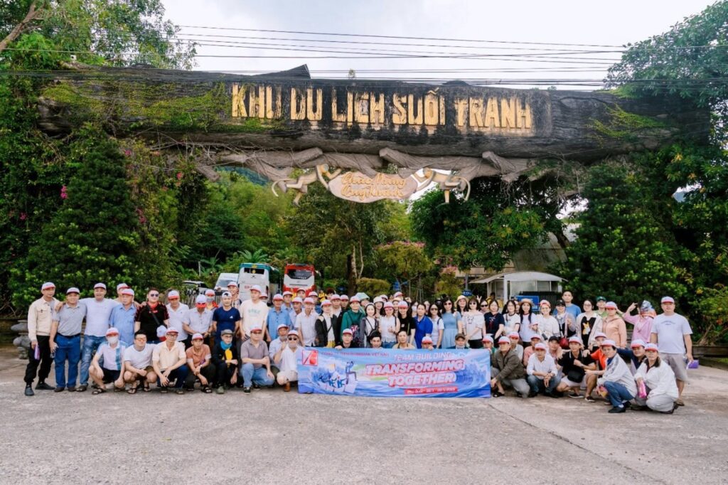 Large group of employees holding a "Transforming Together" banner under the entrance arch of Suoi Tranh Tourist Area.
