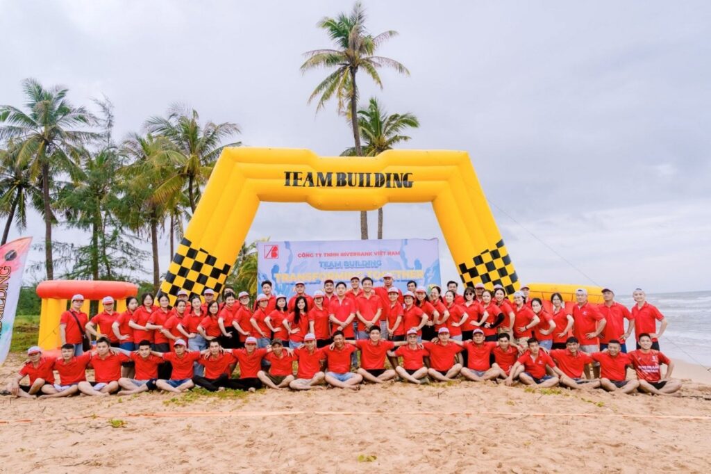 Large group of staff in matching red shirts posing under a yellow inflatable "Team Building" arch on the beach.