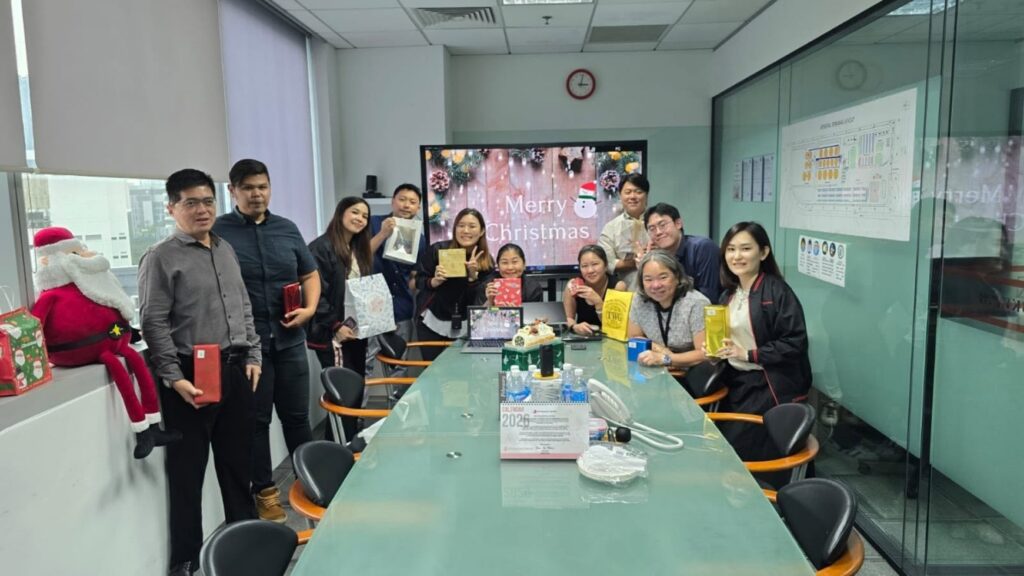 Employees gathered around a meeting room table holding gift boxes during the office Christmas celebration.