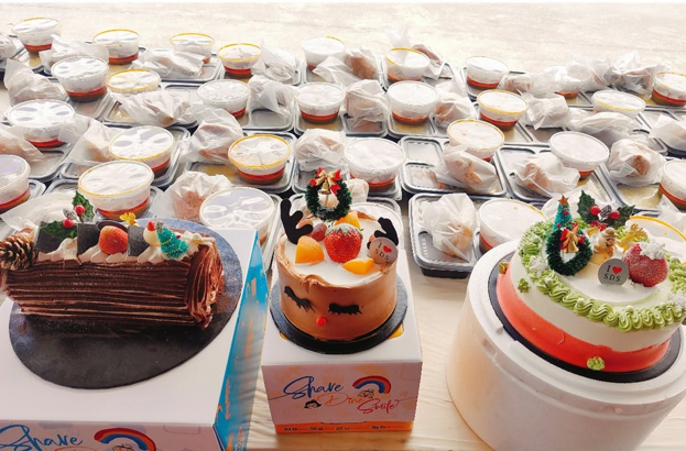Close-up view of three festive Christmas cakes surrounded by rows of packaged meals on a table.