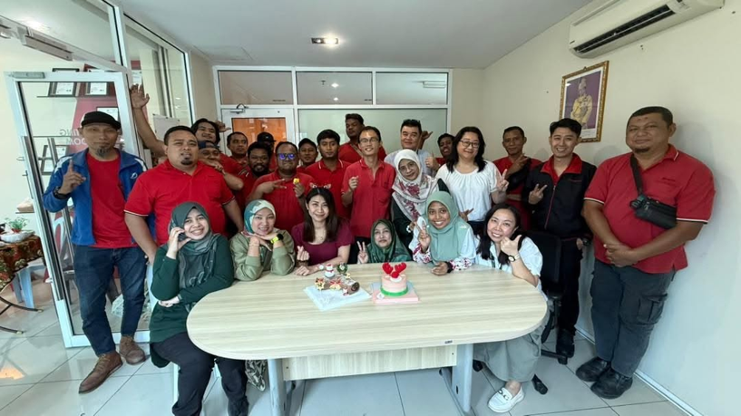 Large group of office employees smiling around a meeting table with small celebration cakes.