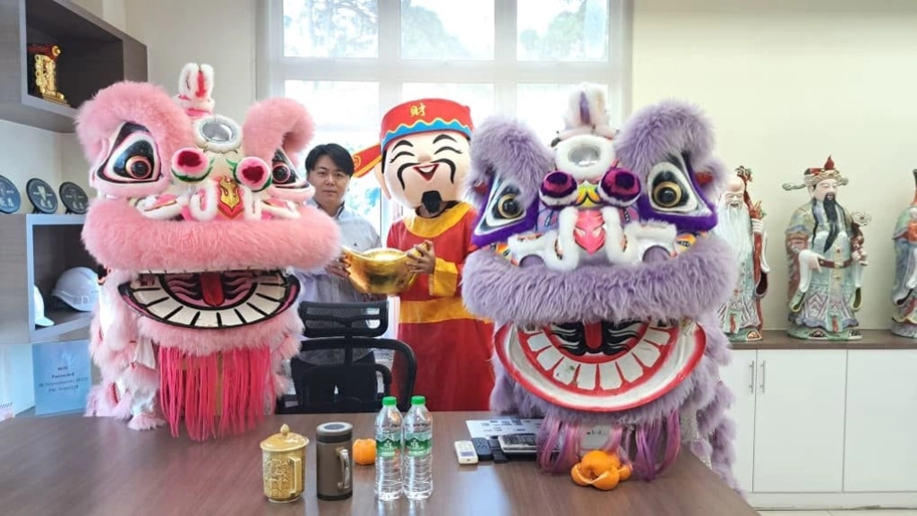 Two people, one in a God of Wealth costume holding a gold ingot, posing indoors between a pink and a purple lion dance head.