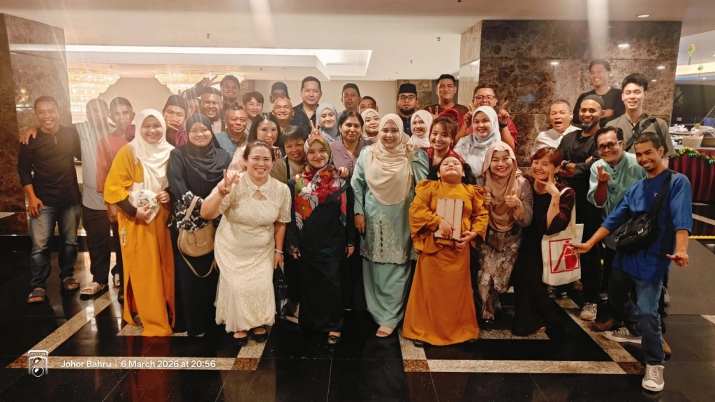 A large group of employees posing for a photo in a brightly lit lobby, with many wearing traditional Malay attire such as baju kurung and baju melayu.
