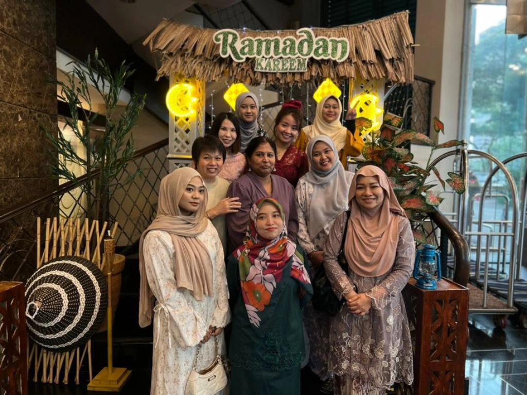 A group of nine women standing on a staircase in front of a rustic, lit-up "Ramadan Kareem" decoration featuring a thatched roof and crescent moons.