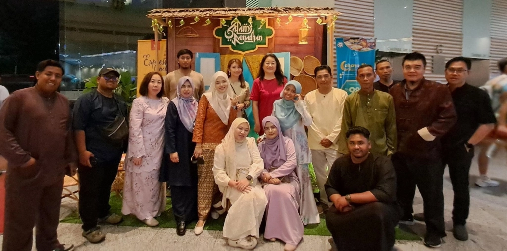 A group of male and female employees posing together in front of a "Salam Ramadhan" photo booth designed to look like a traditional wooden house exterior.