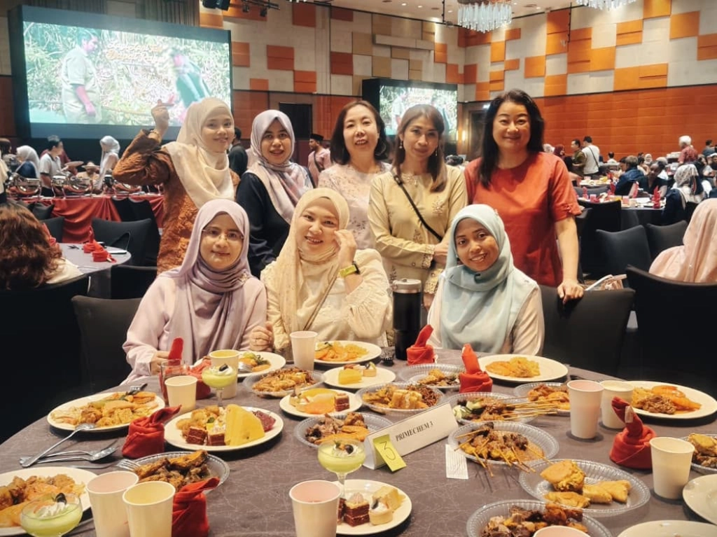A group of eight women posing around a banquet table filled with various dishes during a Ramadan celebration dinner, with a large screen visible in the background.