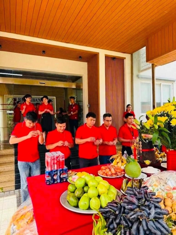 Group of employees in red shirts holding incense and bowing in prayer before a food offering table.