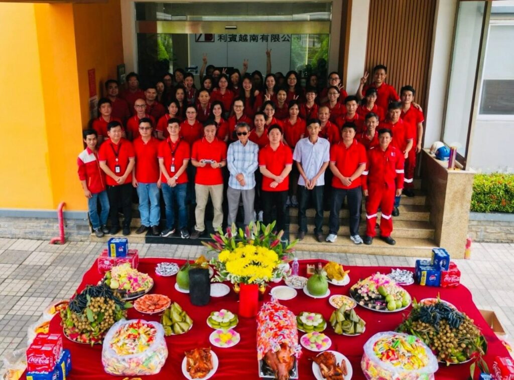 A large group of Riverbank Vietnam employees in red polo shirts posing outside their office behind a table covered in traditional Hungry Ghost Festival food offerings, including a roasted pig, fruits, and flowers.
