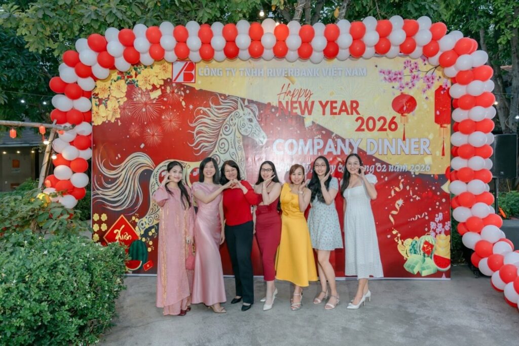 Seven women posing and making heart hand signs in front of a Riverbank Vietnam Happy New Year 2026 Company Dinner backdrop framed by red and white balloons.