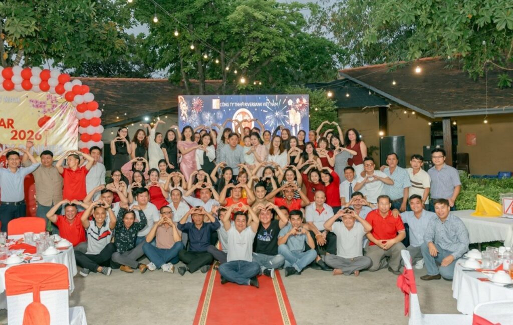 A large group photo of Riverbank Vietnam employees smiling and making heart shapes with their hands during an outdoor company dinner.