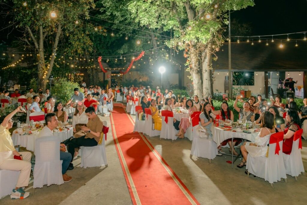 An outdoor evening banquet scene showing attendees seated at round tables with red bows, a central red carpet, and string lights illuminated in the trees.
