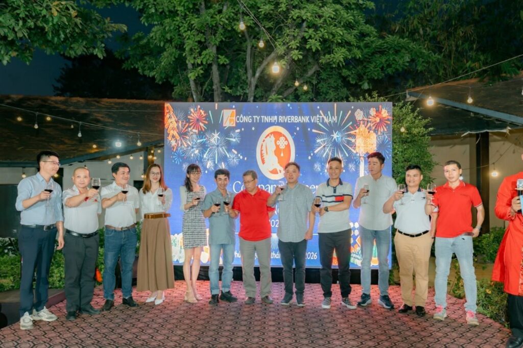 A group of Riverbank Vietnam employees standing on an outdoor stage, toasting with glasses of red wine in front of a digital screen displaying fireworks.