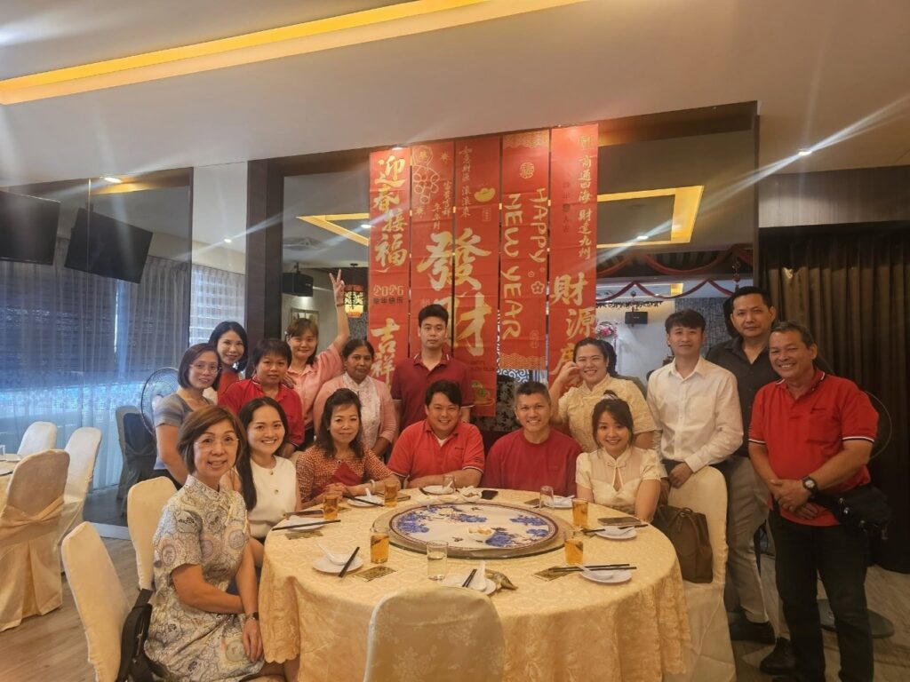 A group of people posing for a photo around a large circular dining table in a restaurant, with red Chinese New Year banners hanging in the background.