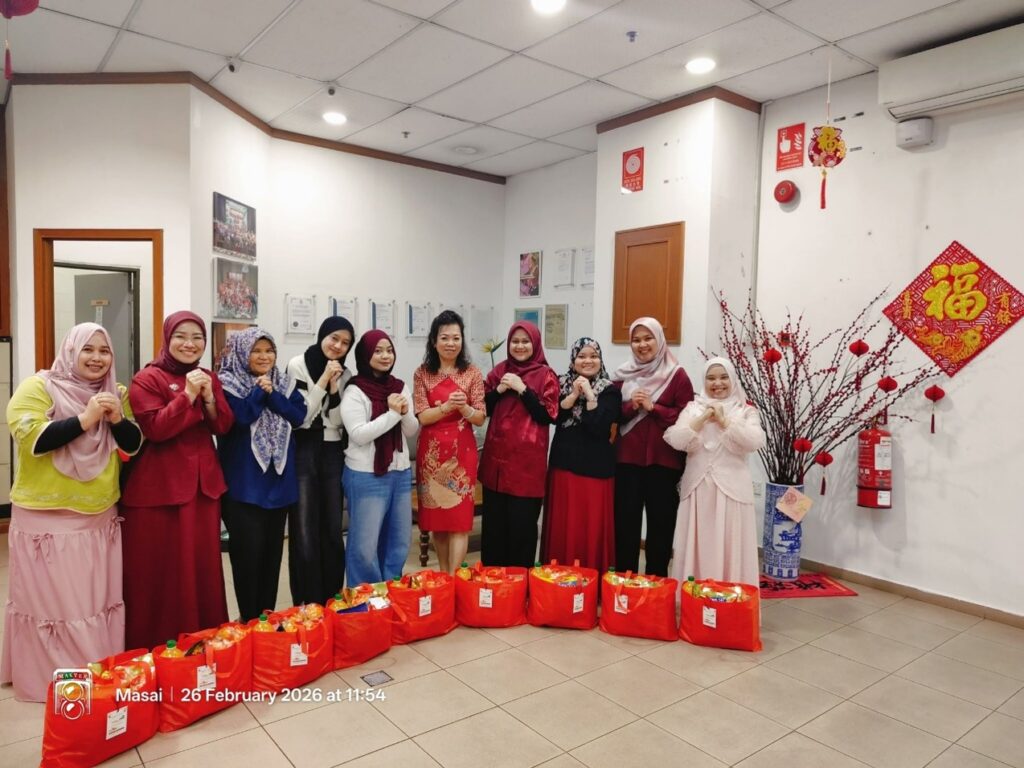 A group of nine women standing indoors giving a traditional Chinese New Year greeting gesture, with a row of red gift bags lined up on the floor in front of them.