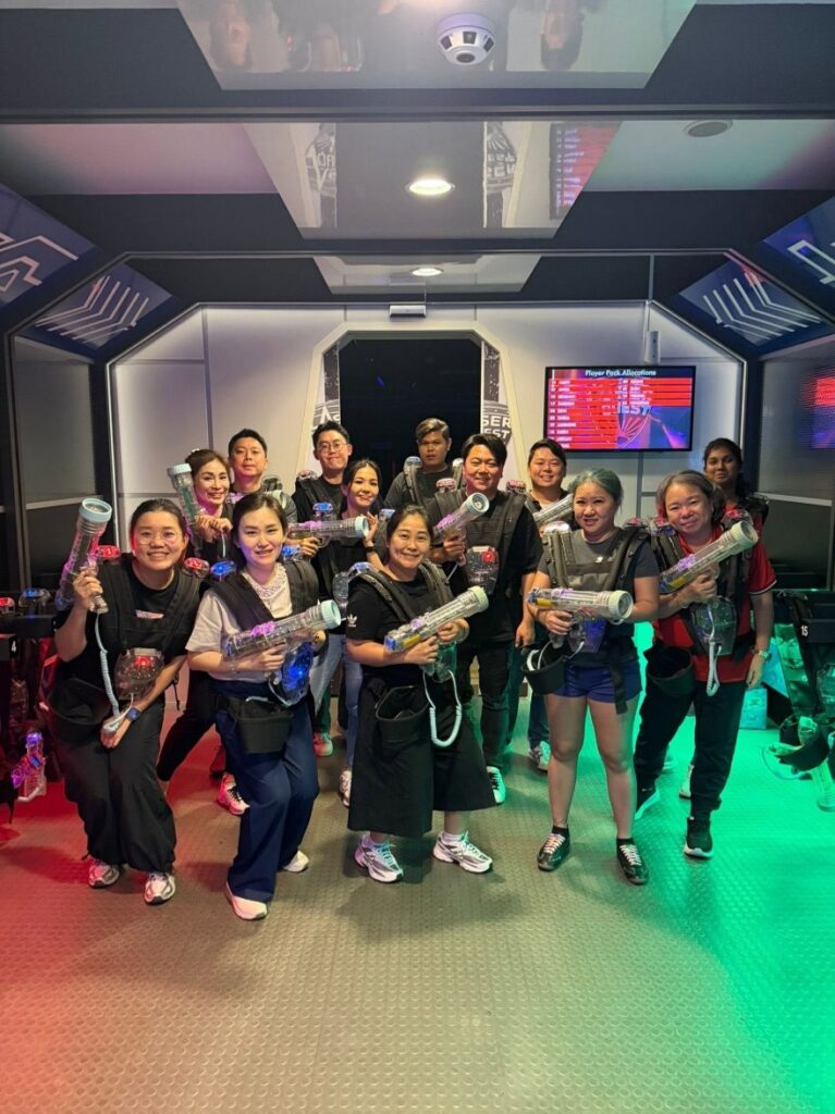 A group of people posing for a photo wearing laser tag vests and holding blasters inside a futuristic, neon-lit laser tag briefing room.
